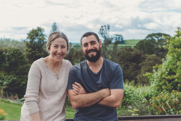 Woman and man in casual clothing smiling with a rural backdrop of green fields behind them.