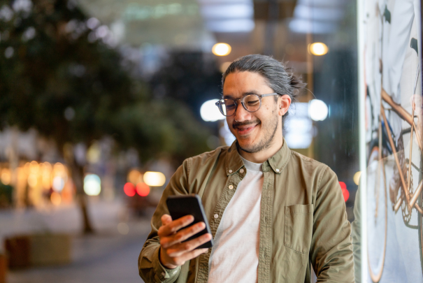 Casually dressed man with dark hair and glasses looking at his mobile phone standing on a city street