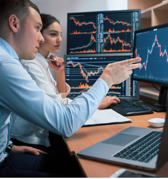 A man and a woman sitting at a computer in an office looking at a graph of the market on a computer to represent active investing | Generate