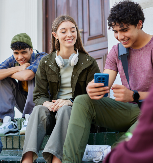 Four teenagers sitting on steps talking and comparing phone screens to represent 16 and 17-year-olds being eligible for KiwiSaver contributions