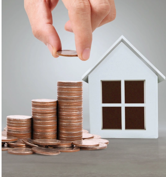A hand adding a coin to a pile of 3 coins with a white toy house in the background to represent New Zealand house prices and the prosperity question
