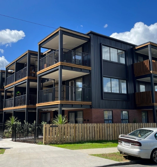 Photo of the Te Puna Hapori housing development - a 3-level building in dark wood showing balconies and courtyards with plants and fencing to represent Generate's investment in the community housing project